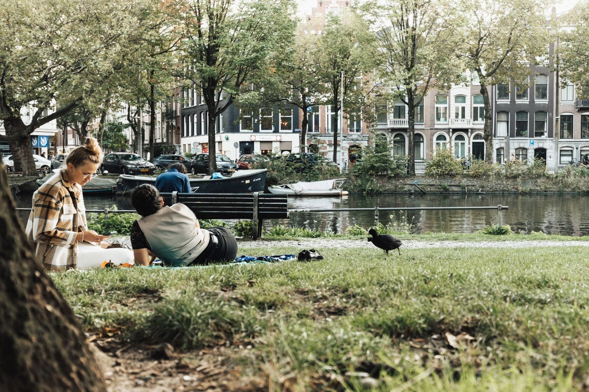 Zwei Personen, die sich im Gras neben einem Amsterdamer Kanal entspannen und picknicken, mit Grachtenhäusern und Booten im Hintergrund.