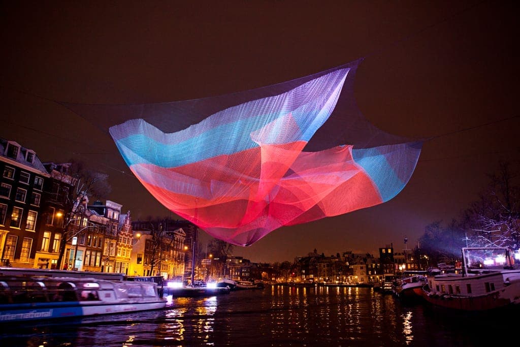 Bunte beleuchtete Netz-Kunstinstallation über einem Kanal in Amsterdam bei Nacht, mit historischen Gebäuden und Touristenbooten während des Light Festival.