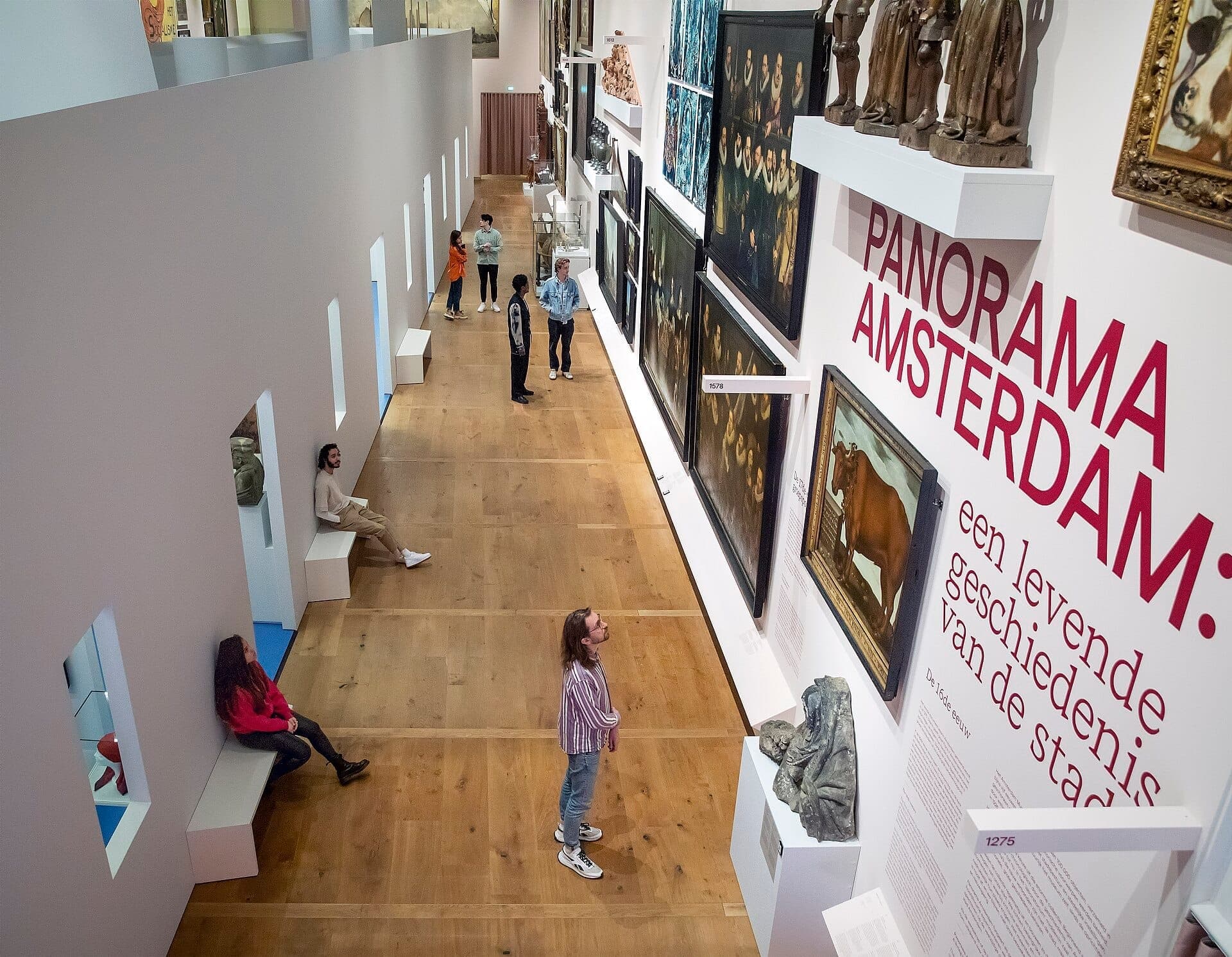 Besucher erkunden die Galerie des Amsterdam Museum und bewundern historische Gemälde und Artefakte, die an weißen Wänden mit Holzboden und modernen Bänken ausgestellt sind.