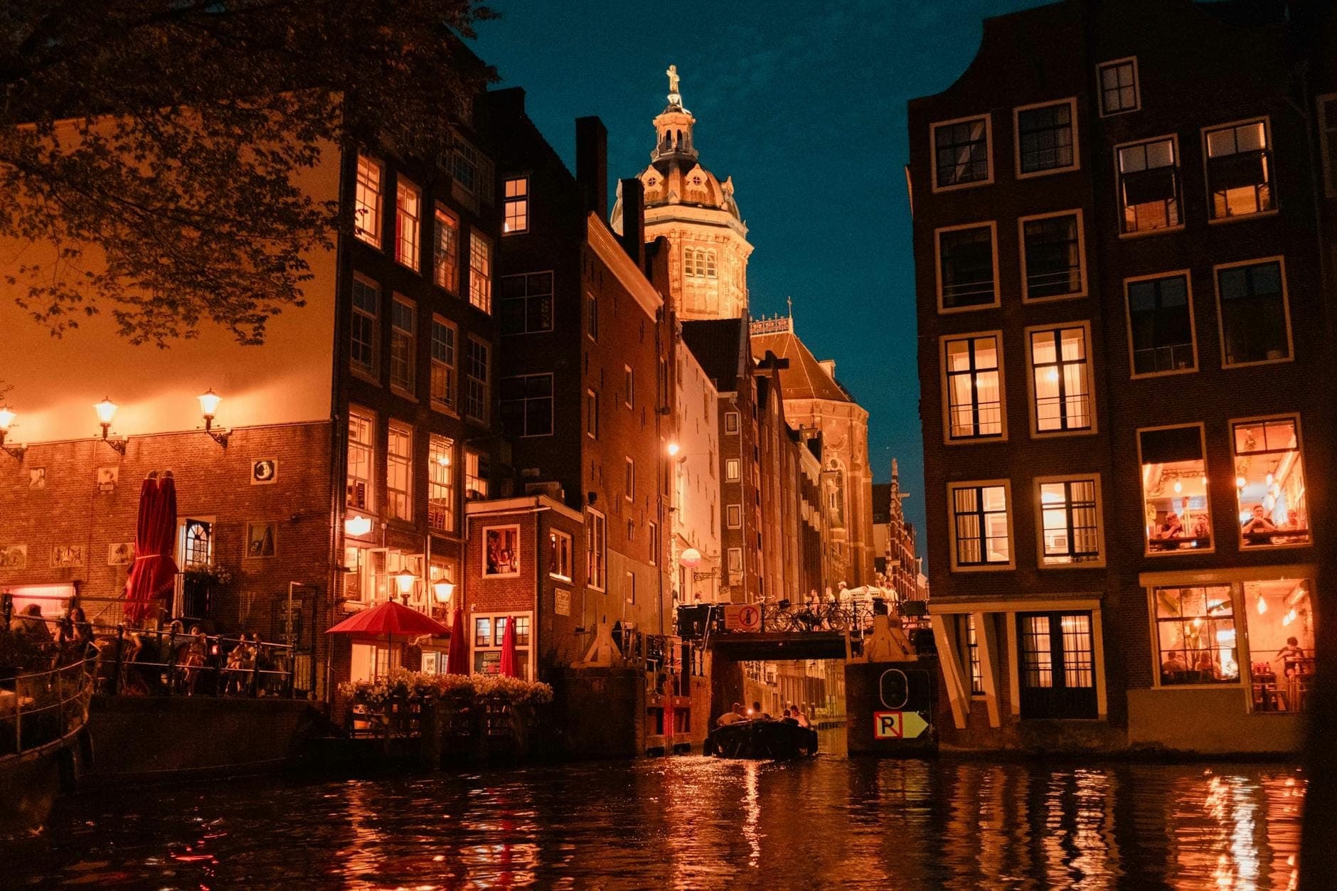 Amsterdamer Kanal bei Nacht mit beleuchteten Bars und Restaurants, im Freien sitzenden Menschen und einem Kirchturm im Hintergrund.