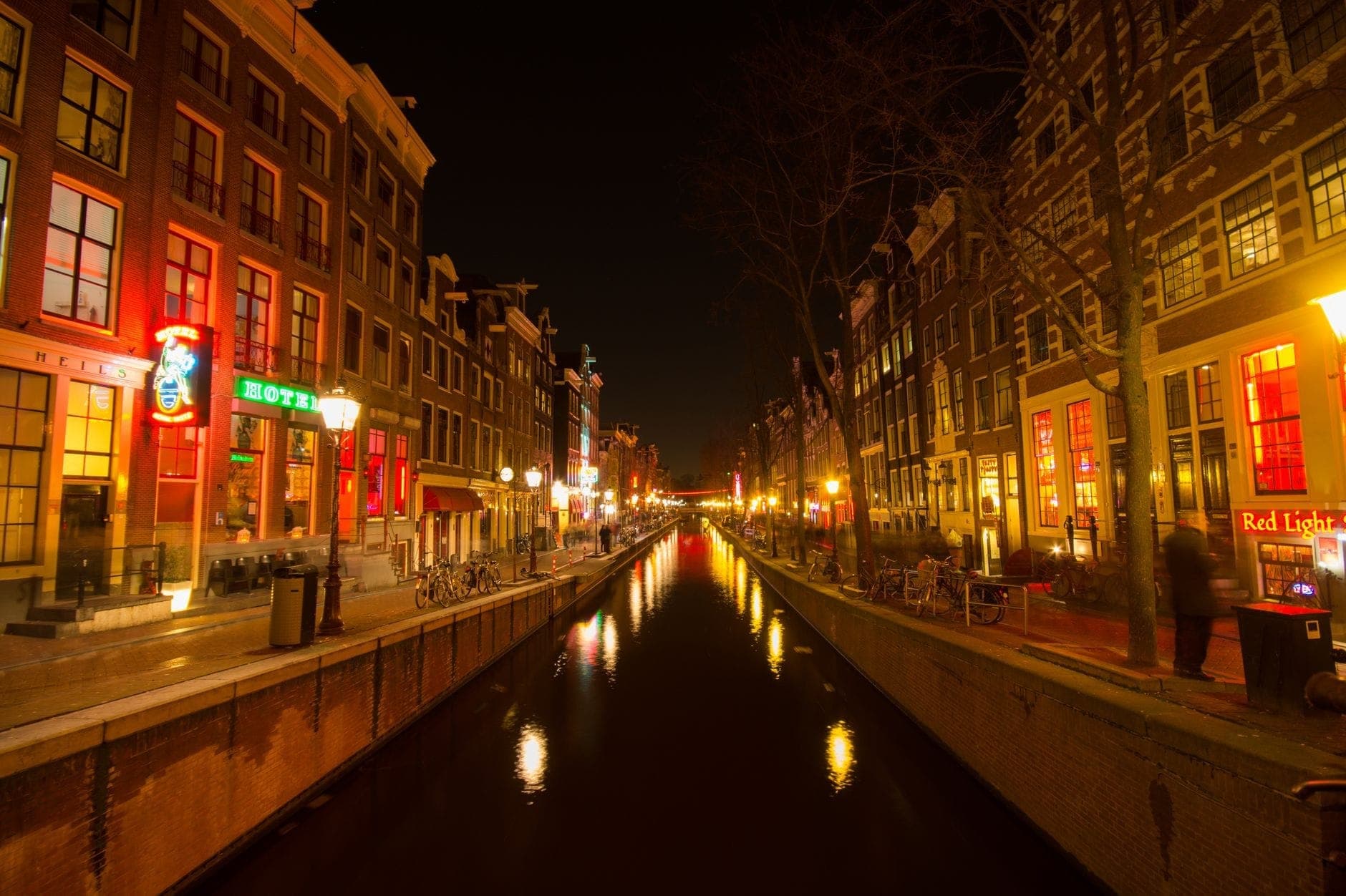Amsterdamer Kanal bei Nacht mit neonbeleuchteten Bars, Hotels und Schildern des Red Light District, die das lebhafte Nachtleben zeigen.