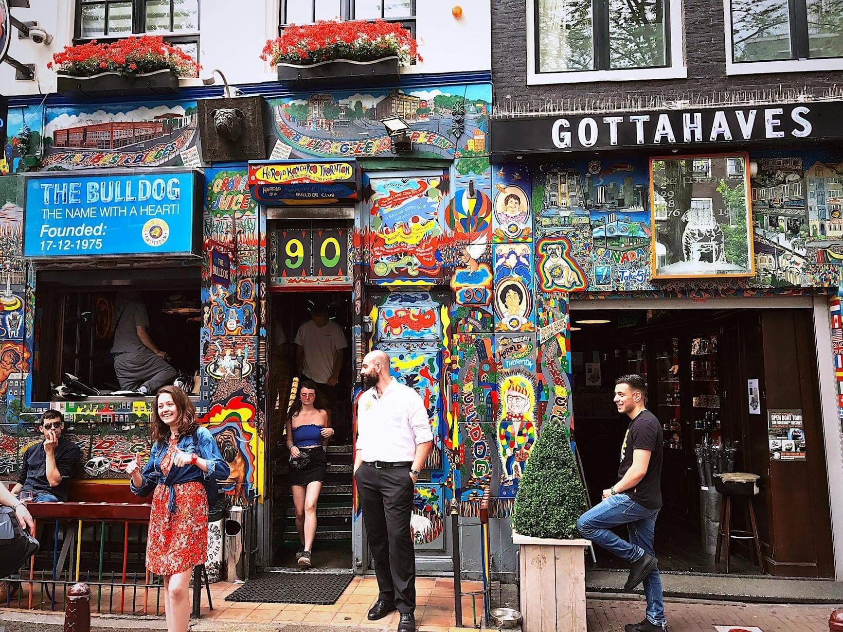 Buntes Außen des Coffeeshops The Bulldog in Amsterdam mit Menschen vor dem Eingang und lebhaften Wandgemälden an der Fassade.