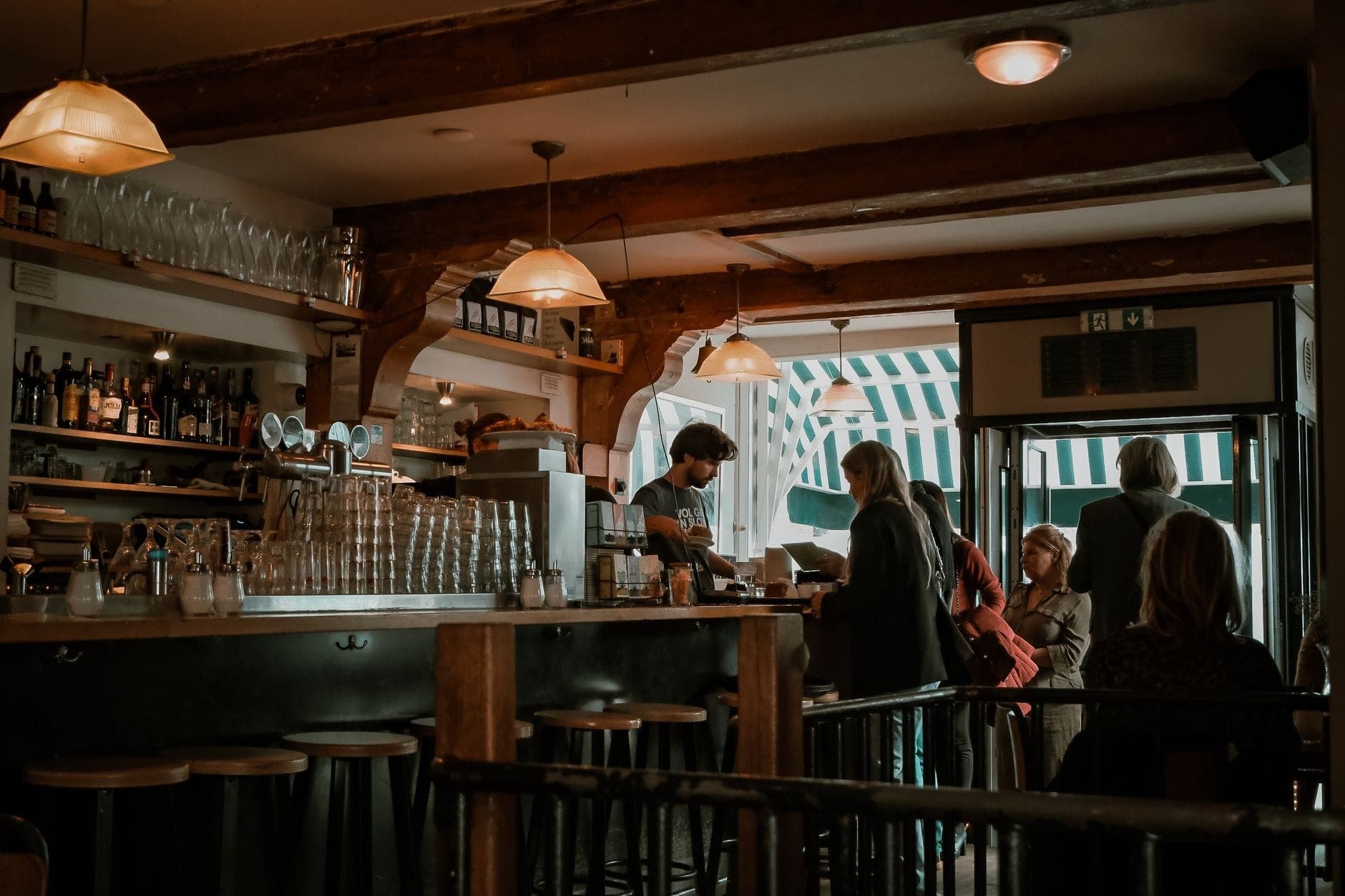 Gemütlich beleuchtetes Barinnere mit Holzbalken, Hockern, Glaswaren und einem Barkeeper, der Gäste bedient, und eine klassische braune Café-Atmosphäre schafft.