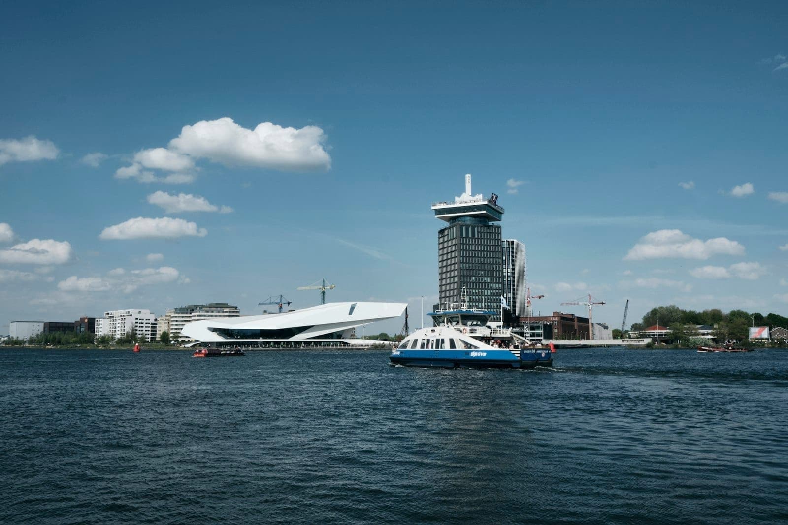 Eine Fähre überquert das IJ mit dem EYE Filmmuseum und dem A'DAM Tower am Ufer von Amsterdam-Noord unter blauem Himmel.