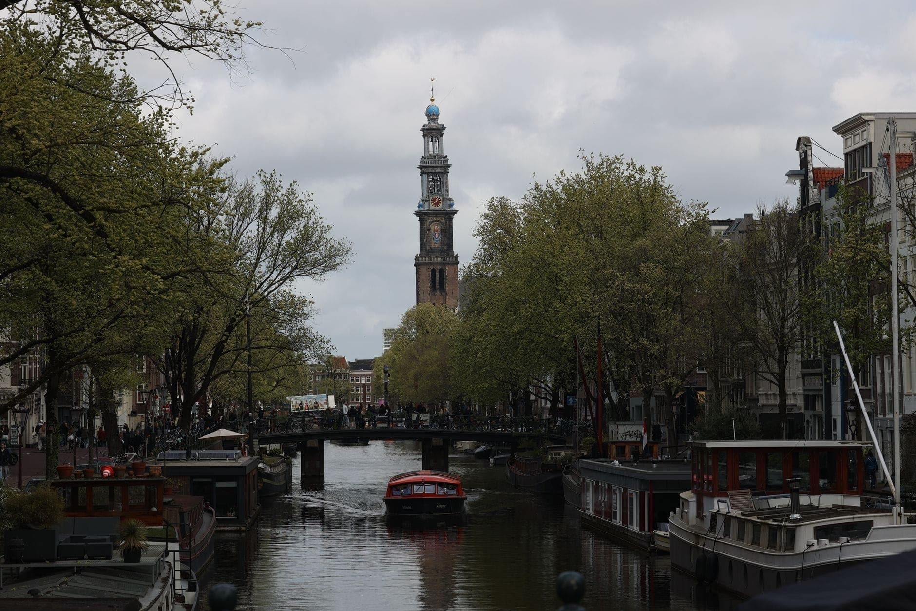 Kanalblick mit Booten und dem Westerkerk-Turm in Amsterdam, flankiert von Bäumen und traditionellen Gebäuden an einem bewölkten Tag.