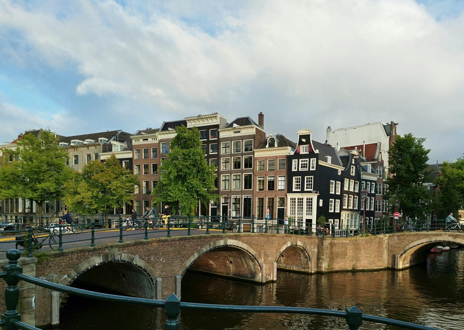 Klassischer Amsterdamer Grachtenblick mit Bogenbrücke, schmalen Giebelhäusern und Bäumen an einem sonnigen Tag – eine Einladung, die Stadt auf eigene Faust zu erkunden.