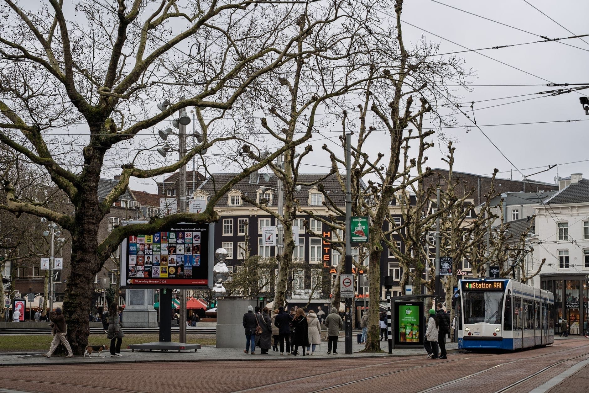 Amsterdamer Straßenbahn an einer Stadthaltetstelle mit wartenden Menschen und Fahrkartenautomaten in der Nähe, die den öffentlichen Nahverkehr in einer realen städtischen Umgebung zeigt.