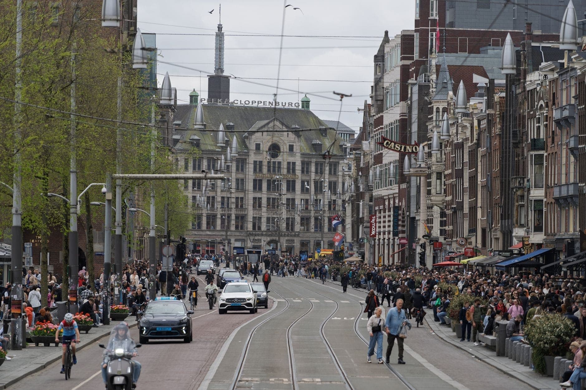 Belebte Stadtstraße in Amsterdam mit Menschenmassen, Radfahrern, Autos und Straßenbahnen an einem bewölkten Tag, die Menschenströme und urbanes Treiben zeigen.