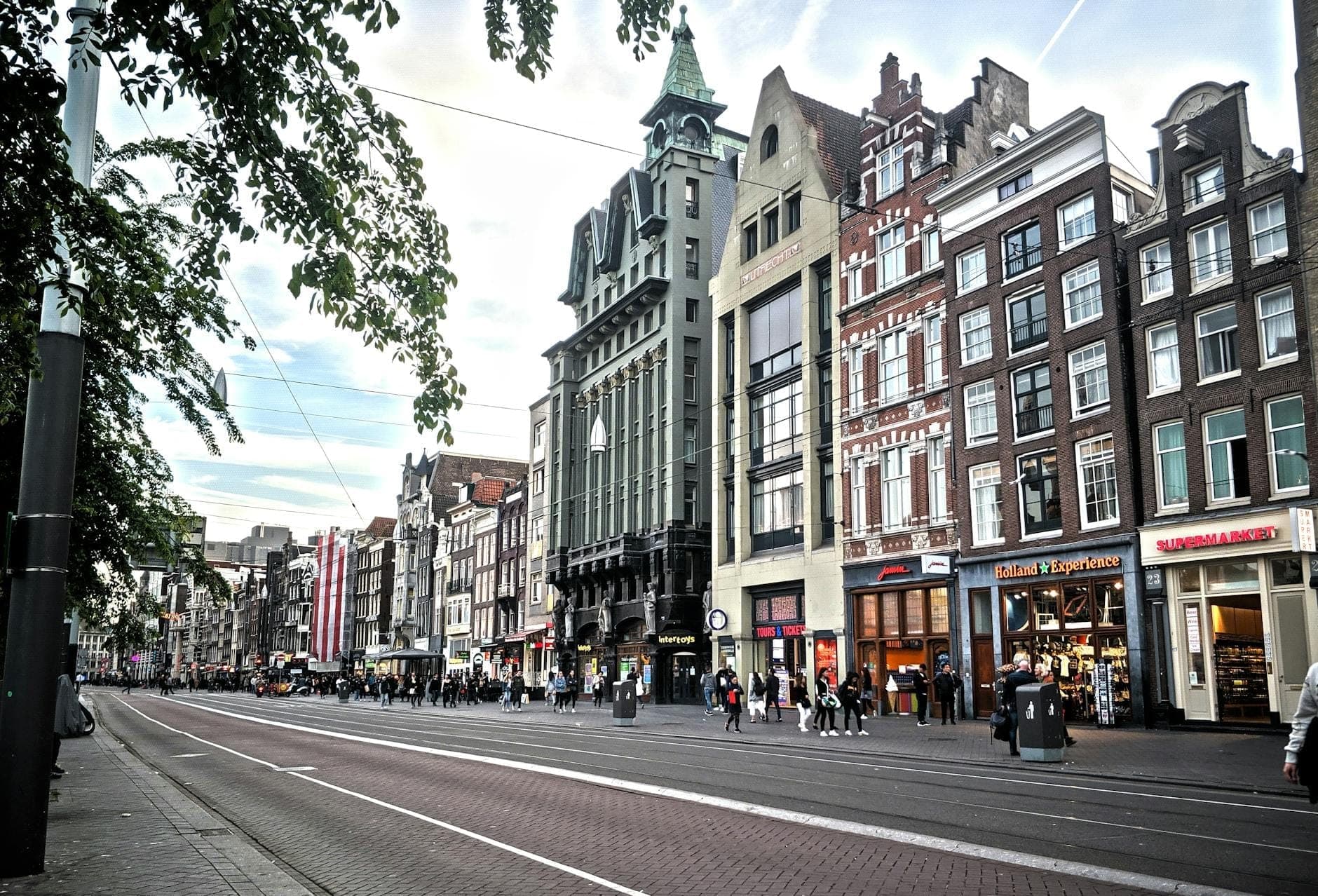 Eine Straße in Amsterdam, gesäumt von historischen Gebäuden und einer Vielzahl belebter Geschäfte, mit sichtbaren Fußgängern auf den Gehwegen und Straßenbahnschienen, die vorbeiführen.