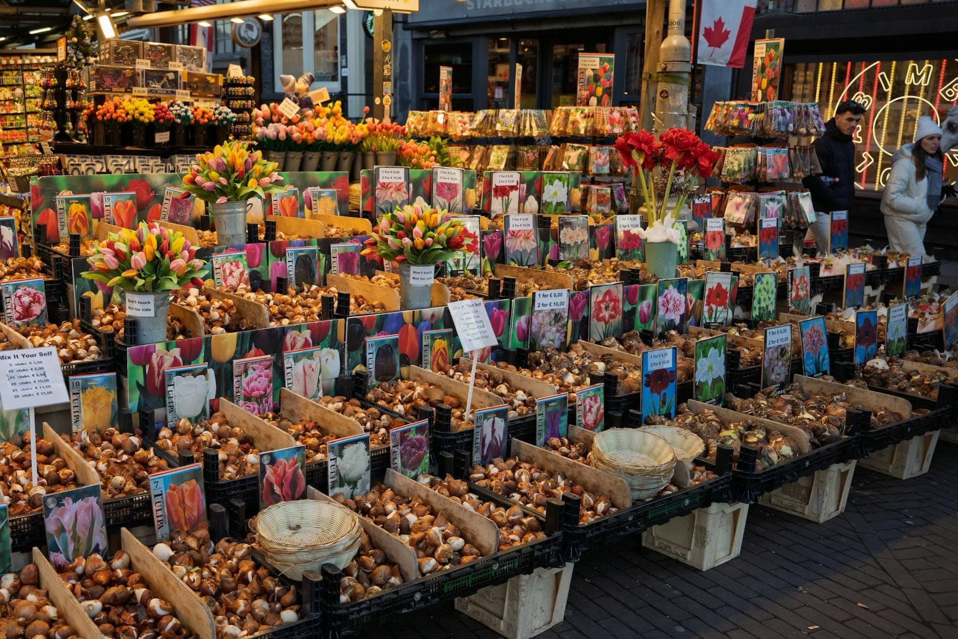 Bunte Blumen- und Zwiebelstände auf einem Freiluftmarkt in Amsterdam mit Einkäufern im Hintergrund.