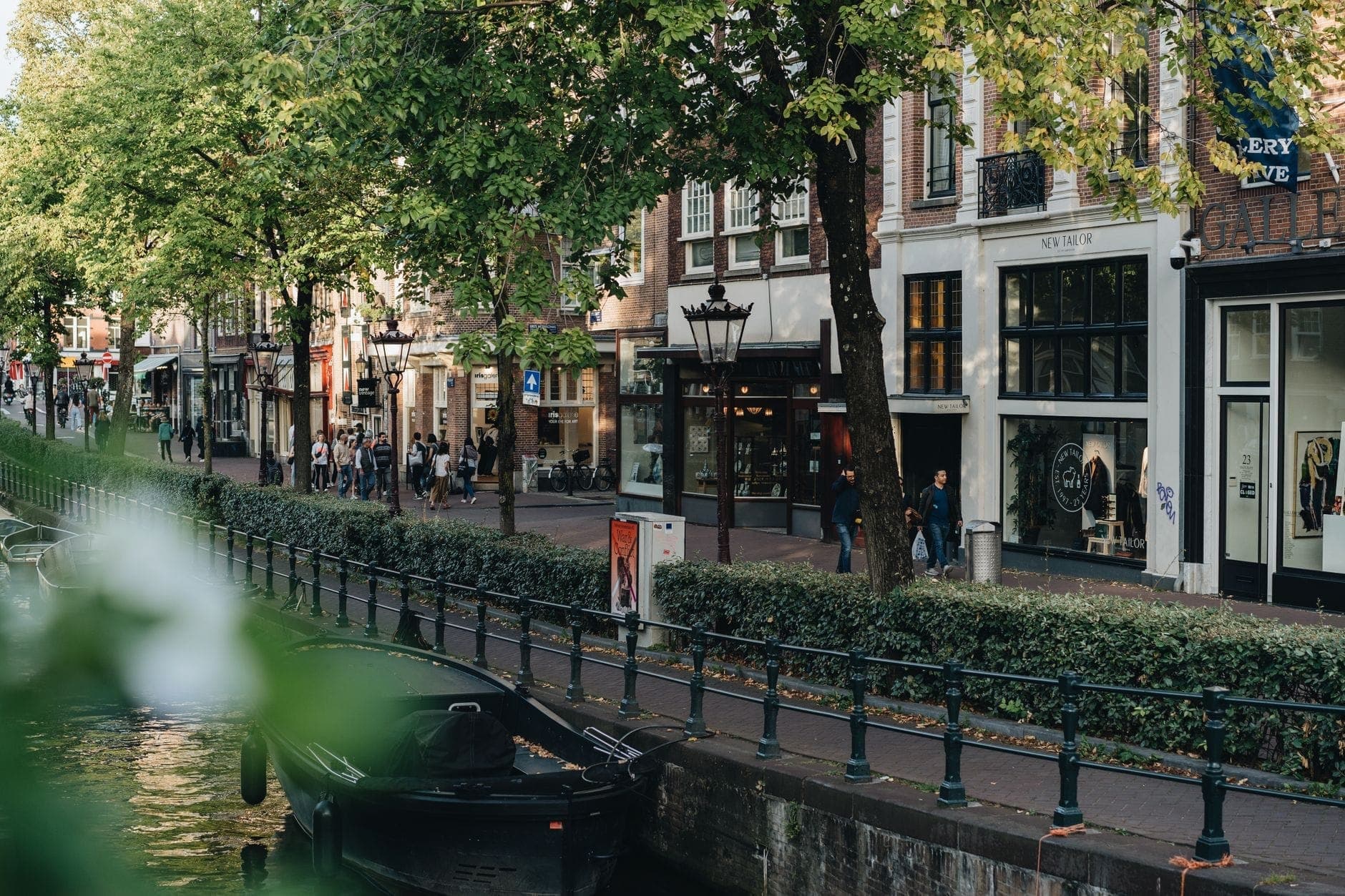 Malerische Einkaufsstraße in Amsterdam entlang eines Kanals, mit Boutiquen, Bäumen und Fußgängern auf dem Gehweg.