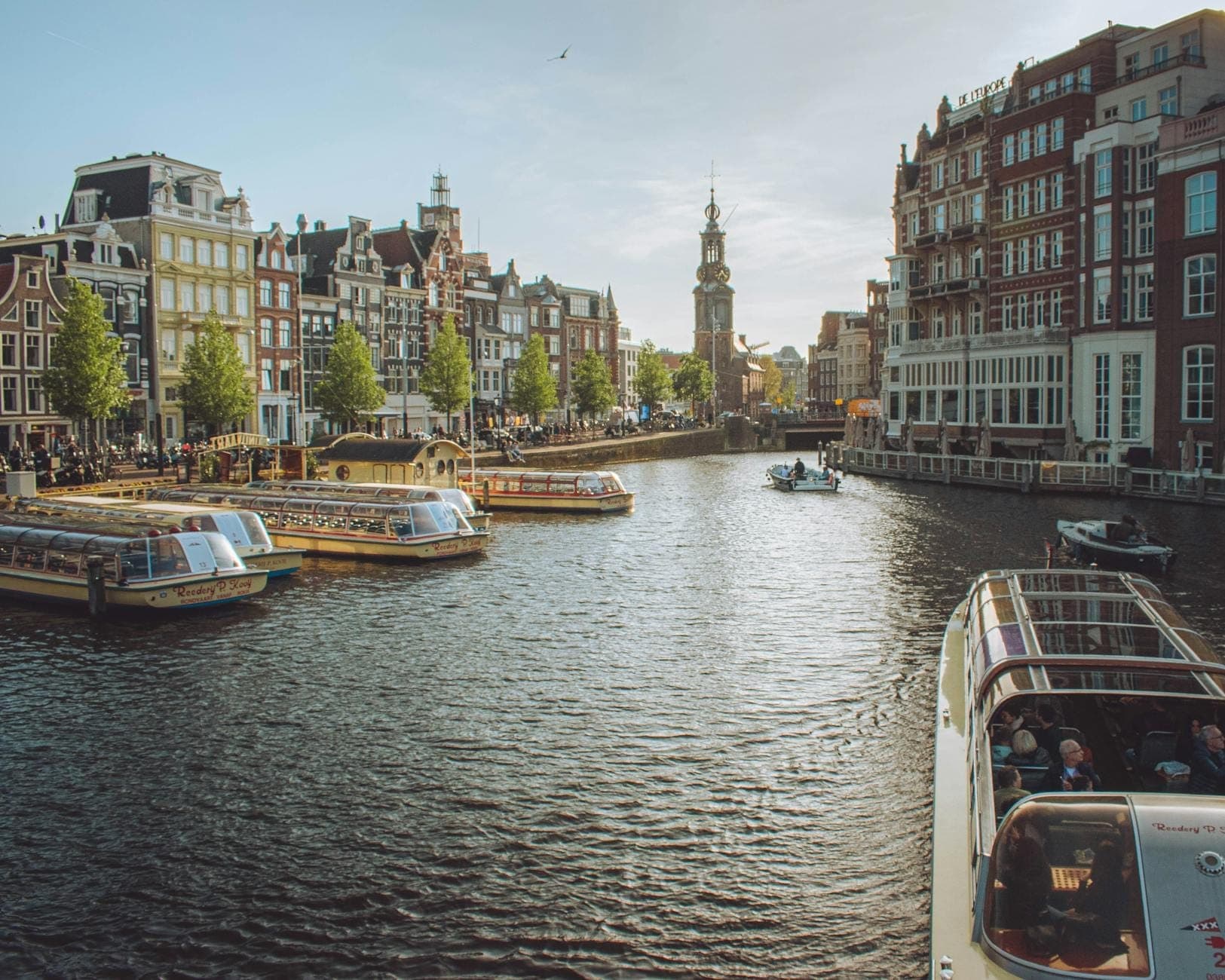 Malerischer Blick auf einen Amsterdamer Kanal mit Ausflugsbooten, historischen Gebäuden, Bäumen und einem Uhrenturm im Hintergrund unter strahlendem Himmel.