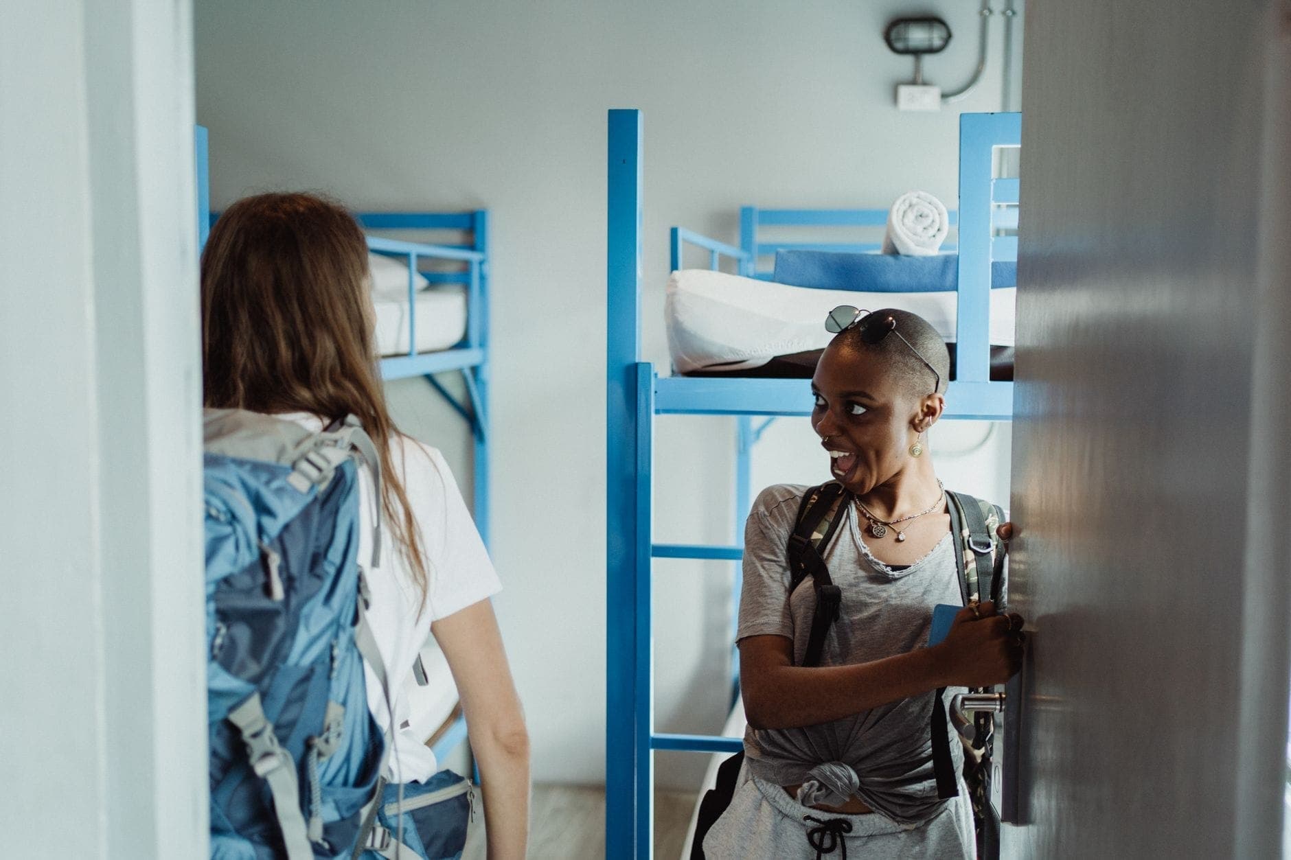 Zwei Alleinreisende mit Rucksäcken betreten ein modernes Hostel-Schlafsaalzimmer mit blauen Etagenbetten und machen sich bereit, sich einzurichten.