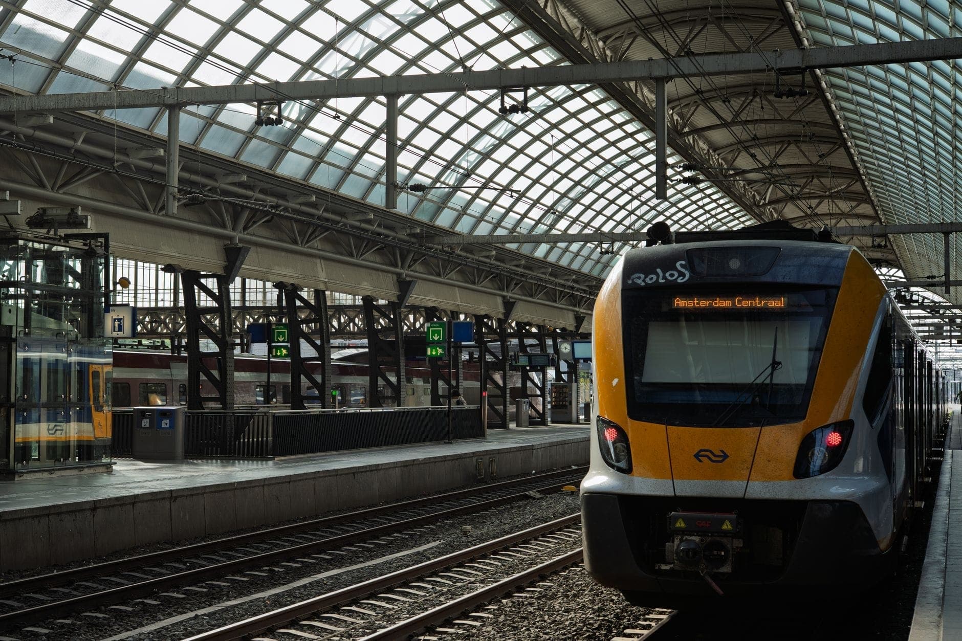 Moderner Zug an einem Bahnsteig im Bahnhof Amsterdam Centraal, unter einem gläsernen Bogendach mit sichtbarer Beschilderung.