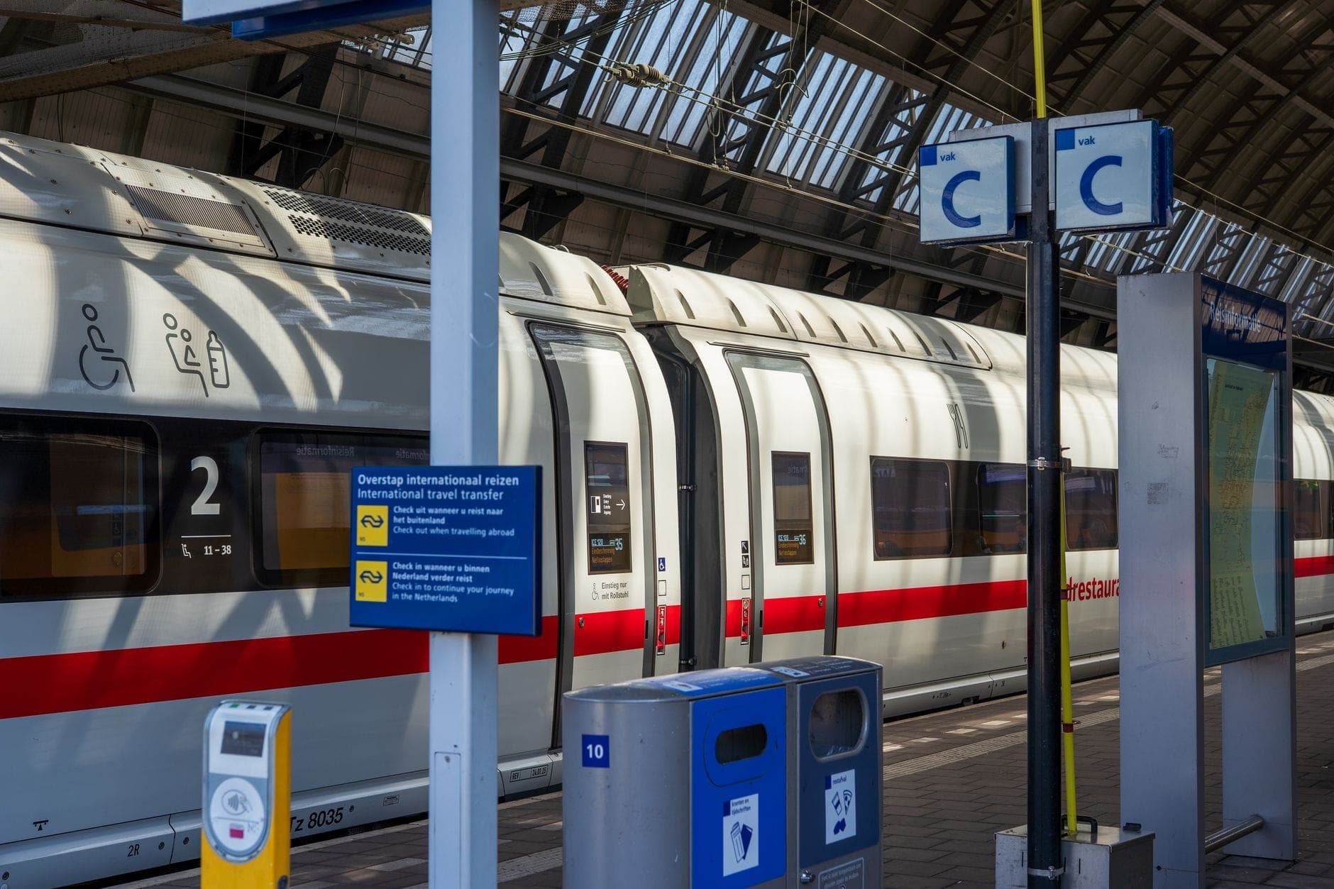 Seitenansicht eines modernen weißen ICE-Zuges mit rotem Streifen an einem Bahnsteig im Amsterdam Centraal, unter einem großen Glasdach.