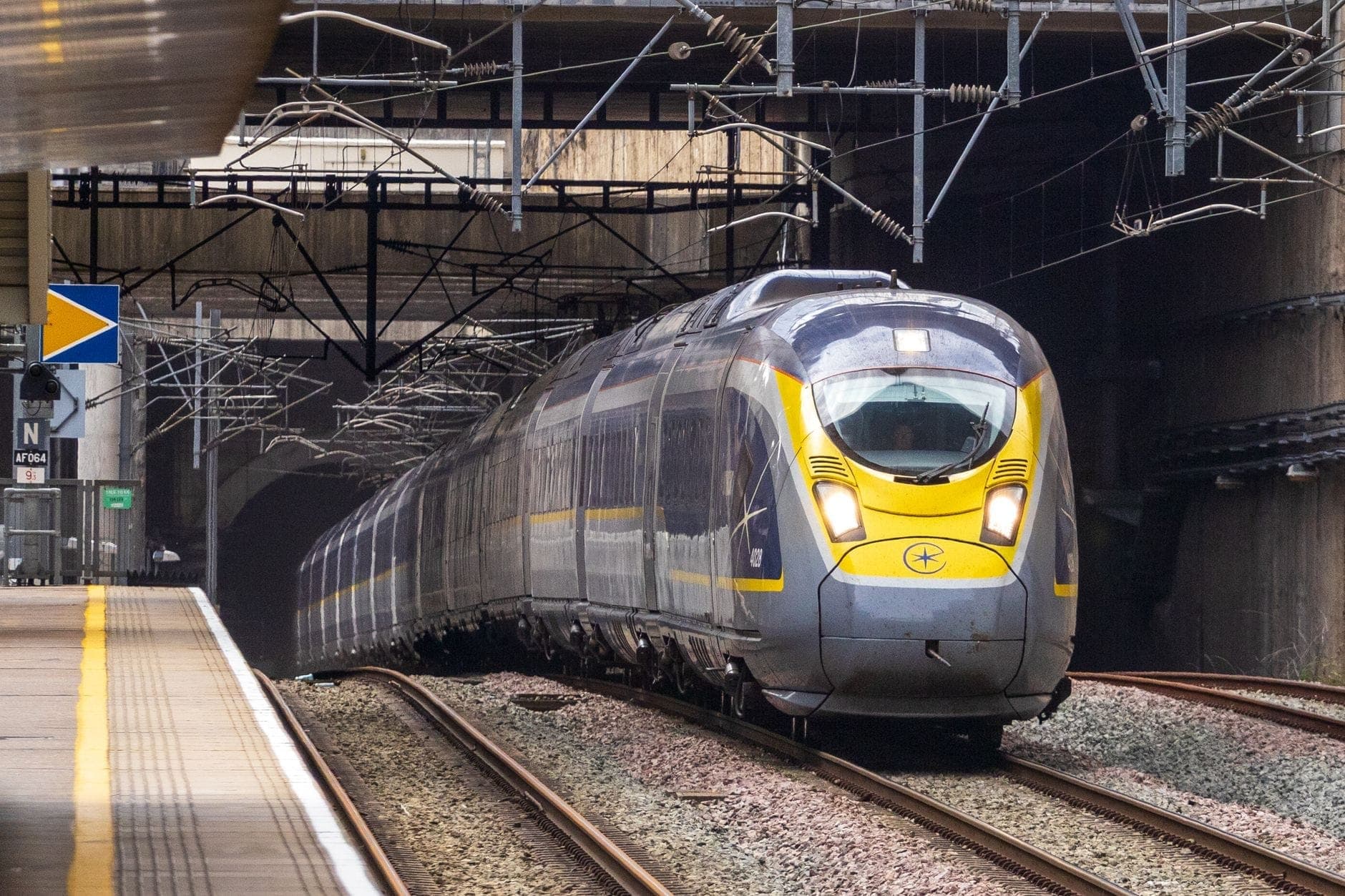 Ein moderner Eurostar-Hochgeschwindigkeitszug, der aus einem Tunnel fährt, mit einem Bahnsteig links und Oberleitungen im Bild.