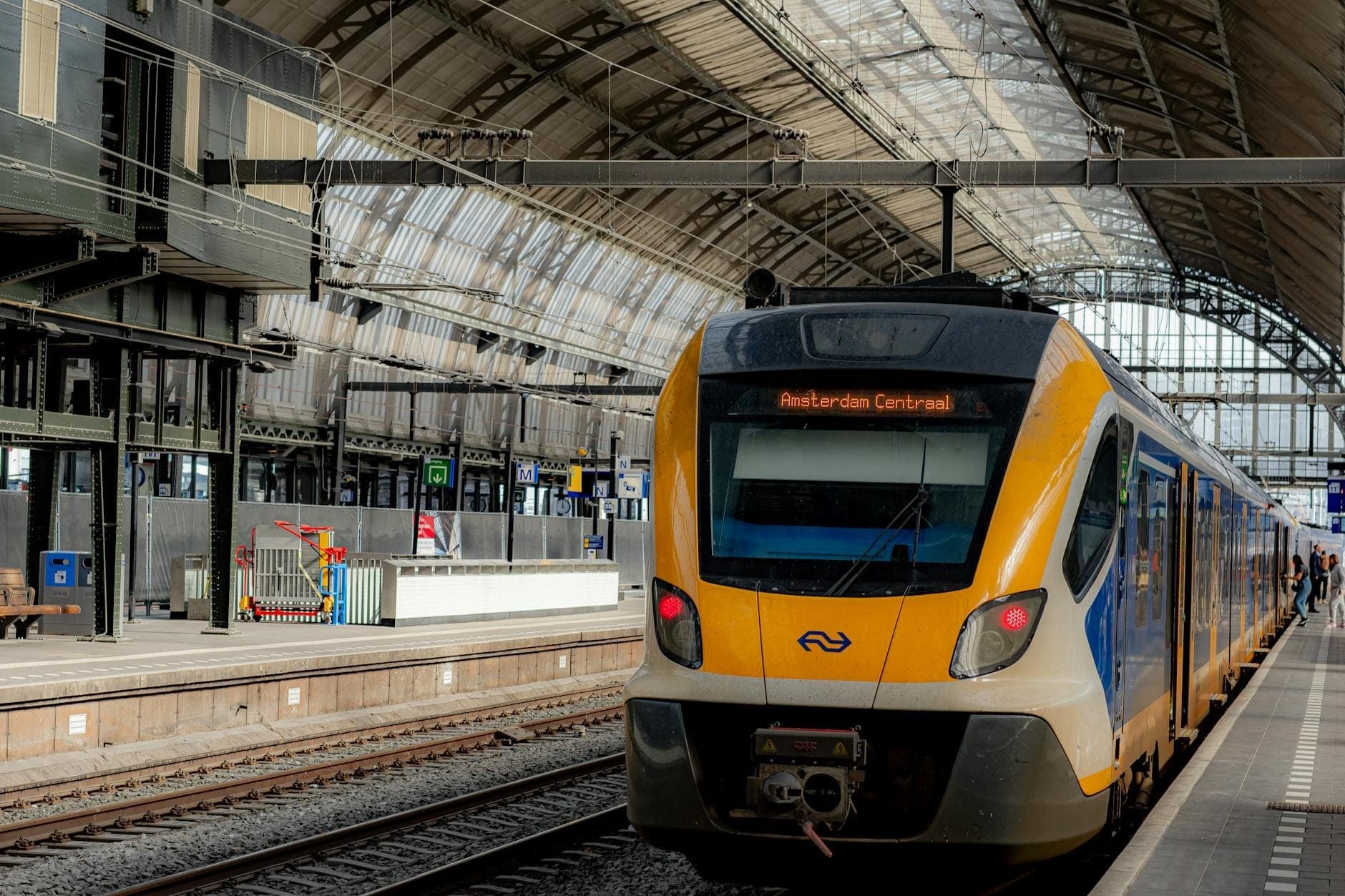 Gelb-blauer niederländischer Zug an einem Bahnsteig im Inneren des Bahnhofs Amsterdam Centraal, unter dem markanten Bogenglasdach des Bahnhofs.