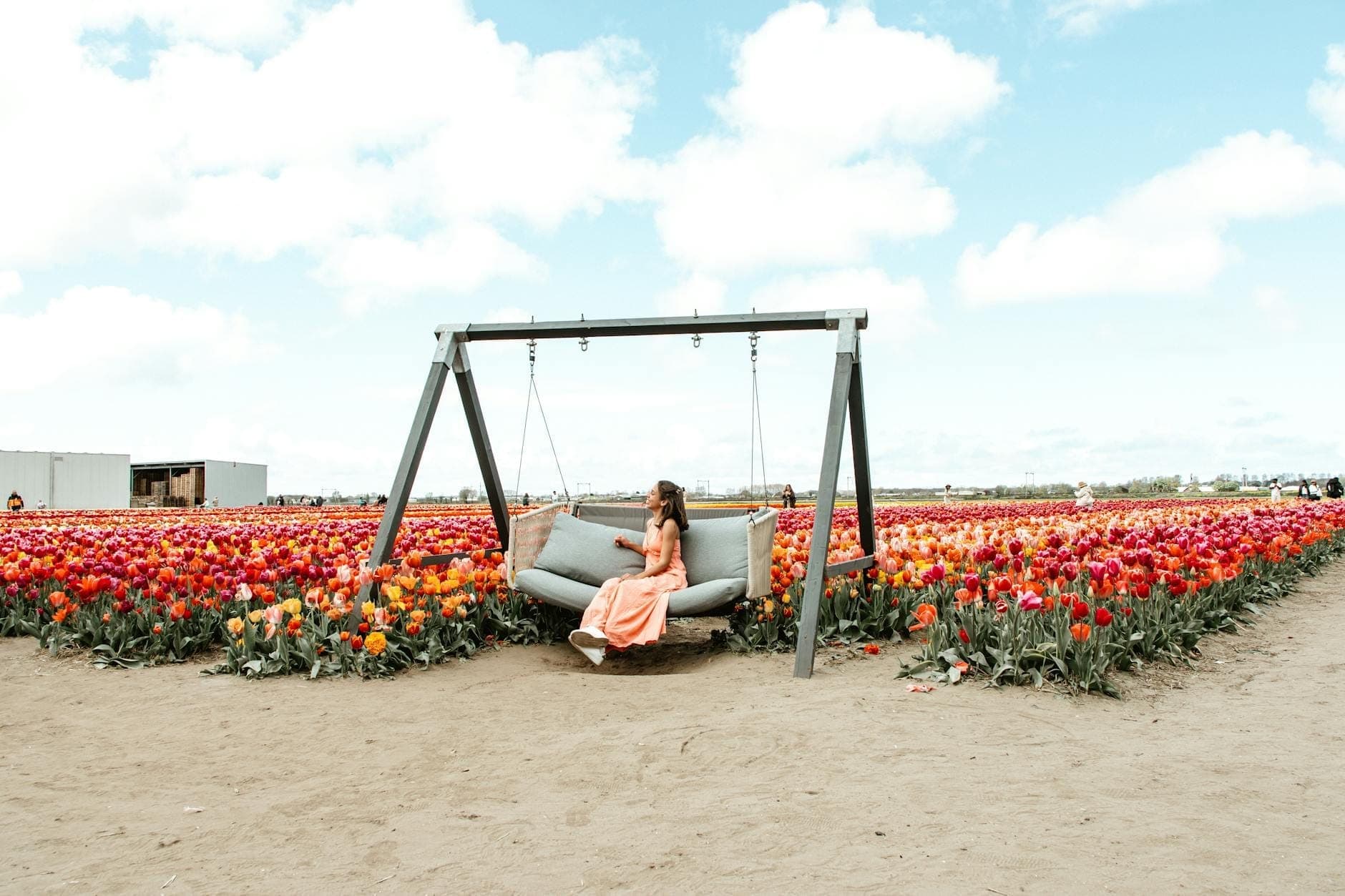 Eine Frau in einem pfirsichfarbenen Kleid sitzt auf einer Schaukel, umgeben von leuchtenden Tulpenfeldern unter blauem Himmel – ein Bild für die Tulpensaison in Amsterdam.