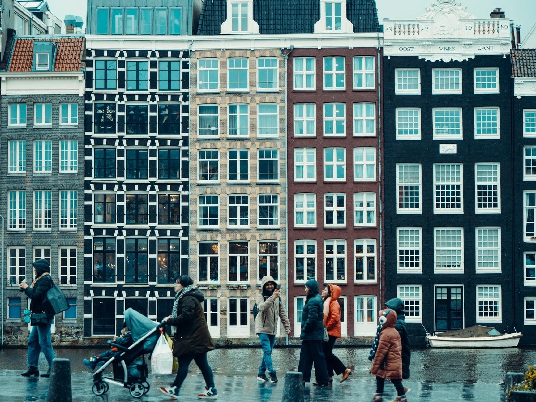 Menschen spazieren vor klassischen schmalen Grachtenhäusern in Amsterdam, mit bunten Fassaden und Spiegelungen auf dem nassen Pflaster – ein Sinnbild für den urbanen Charme der Stadt.