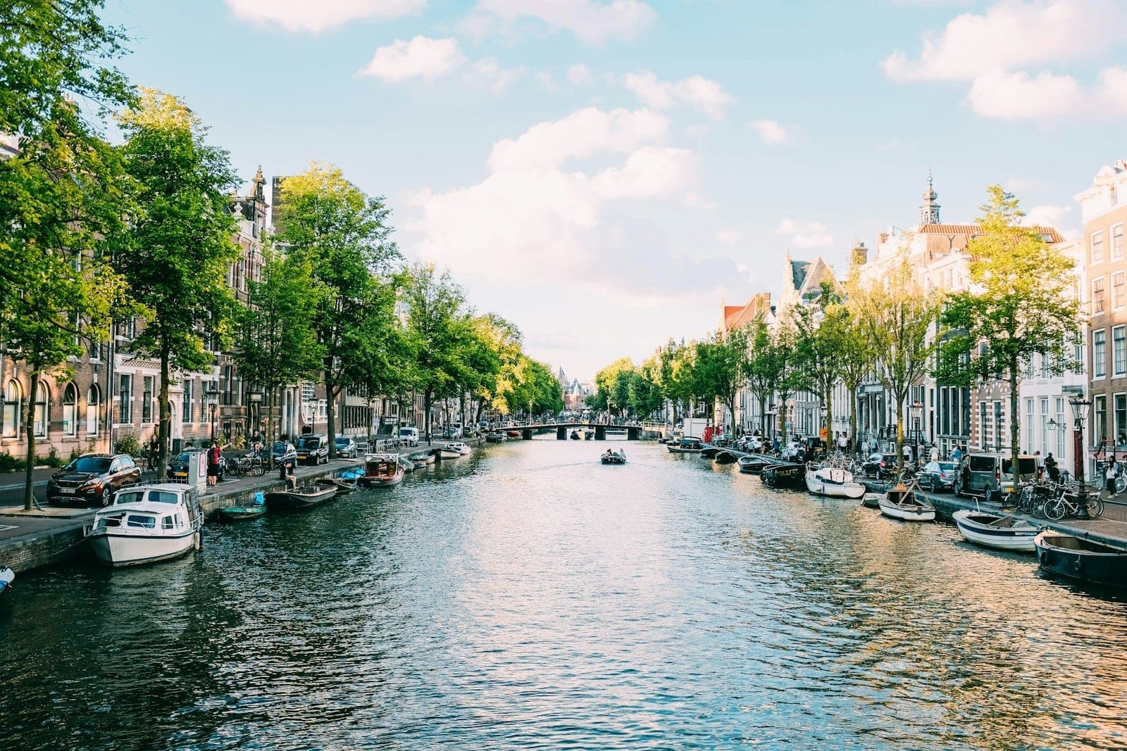 Wide view of an Amsterdam canal lined with traditional Dutch houses, trees, and boats on a sunny day with blue sky and clouds.
