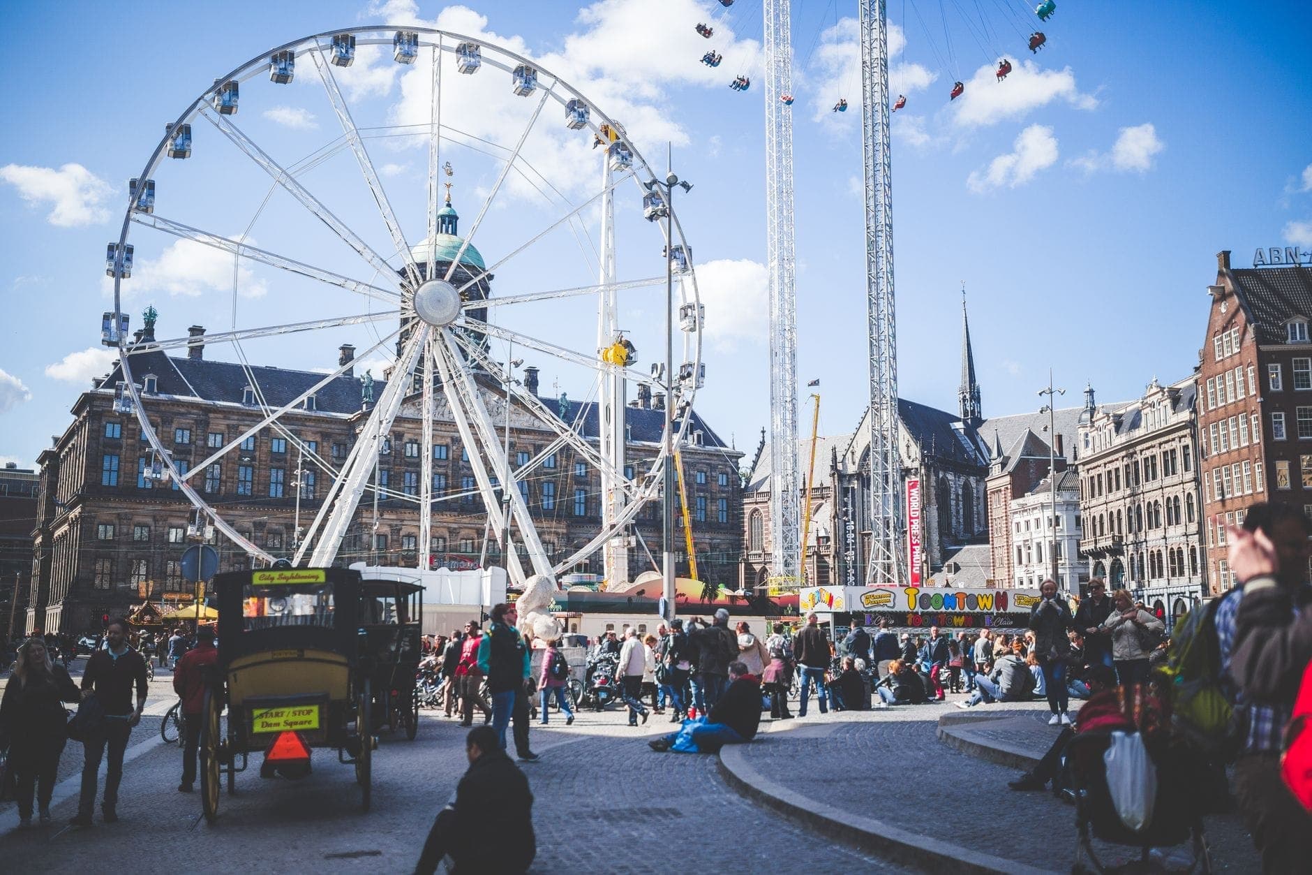 Belebter Stadtplatz in Amsterdam mit einem großen Riesenrad, historischen Gebäuden und vielen Familien und Kindern, die einen schönen Tag unter strahlend blauem Himmel genießen.