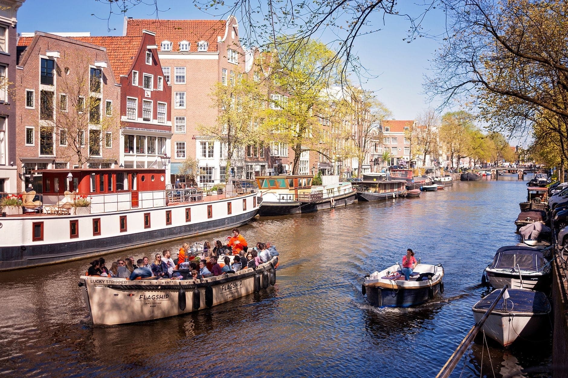 Offenes Rundfahrtboot mit Menschen, die eine Fahrt auf einem Amsterdamer Kanal genießen, umgeben von historischen Häusern und anderen Booten an einem sonnigen Tag.