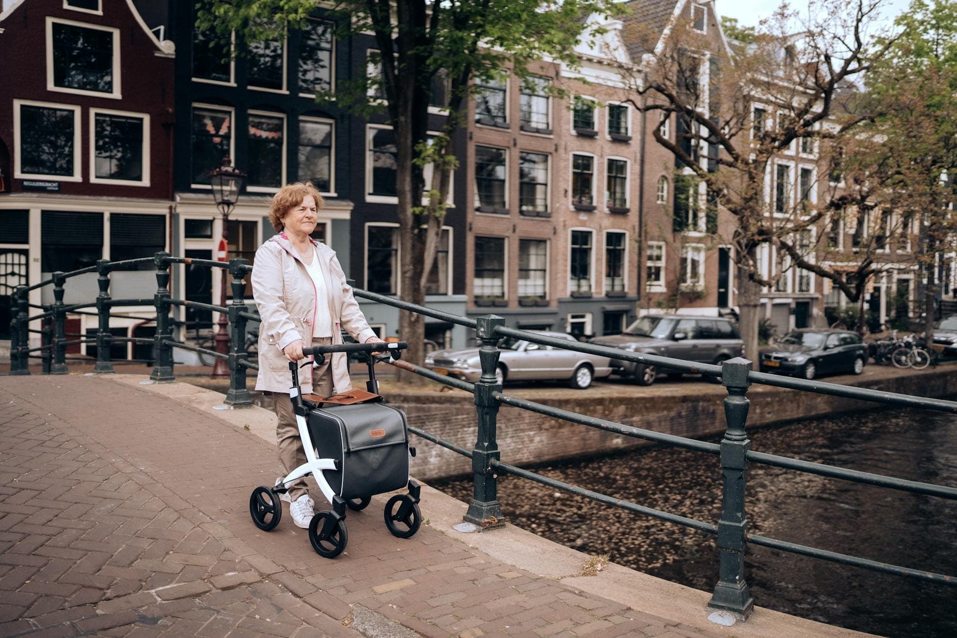 Frau mit Rollator überquert eine Brücke über eine Gracht in Amsterdam, mit klassischen Grachtenhäusern und Fahrrädern im Hintergrund.