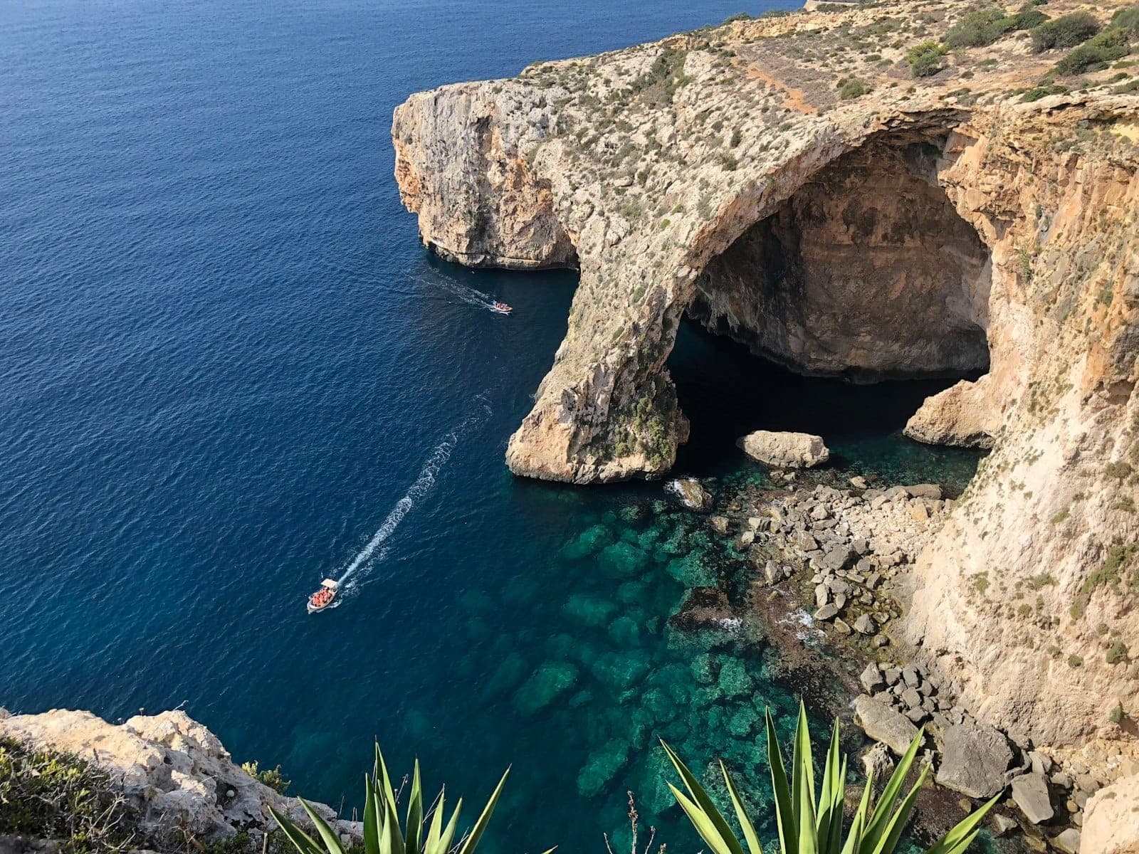 Vista aerea delle scogliere e dell'arco della Blue Grotto di Malta con acqua blu brillante, barche e vegetazione costiera.