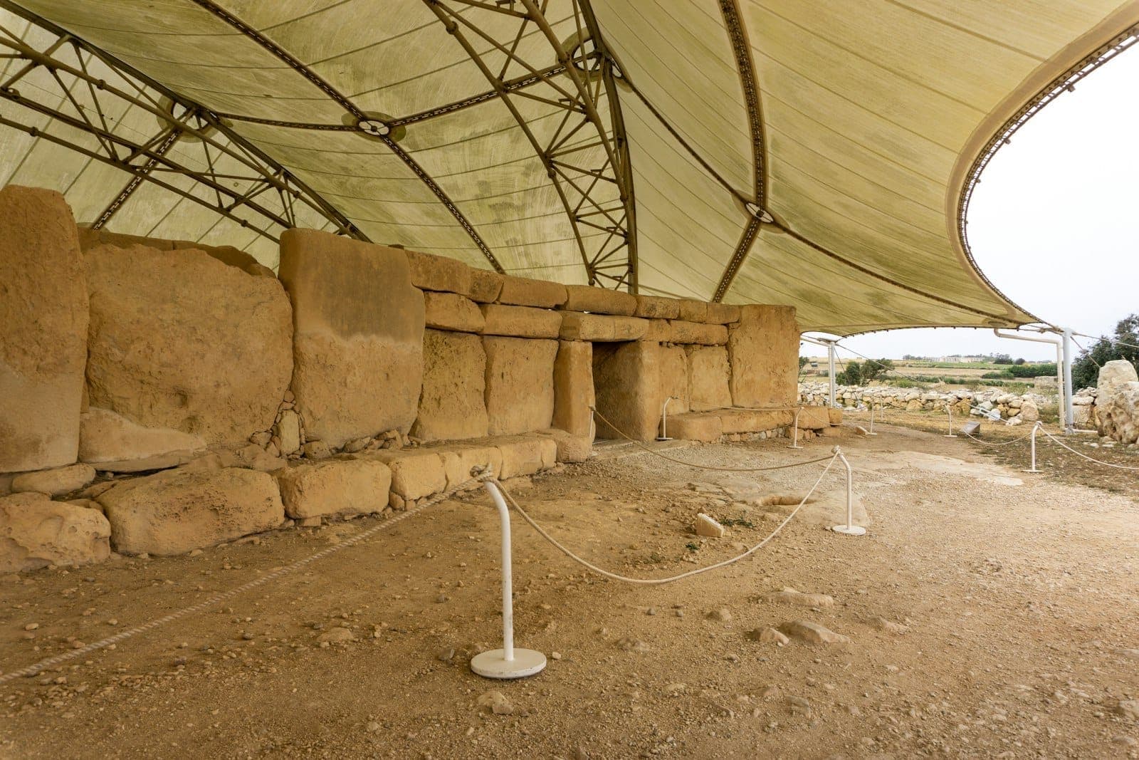 Rovine di un antico tempio in pietra sotto una moderna struttura di copertura protettiva, con percorsi delimitati da corde in un sito archeologico.