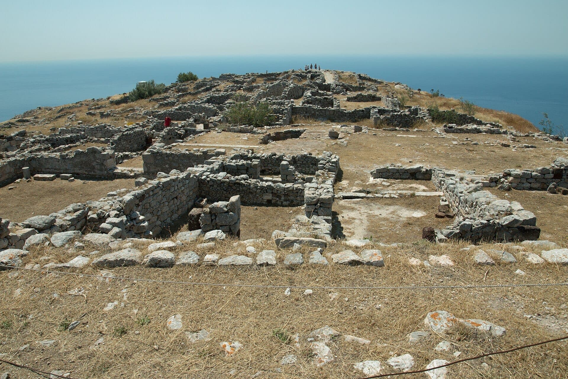 Steinruinen des antiken Thera auf einem sonnenbeschienenen Plateau, mit Blick auf das tiefblaue Ägäische Meer am Mesa-Vouno-Kamm auf Santorini.