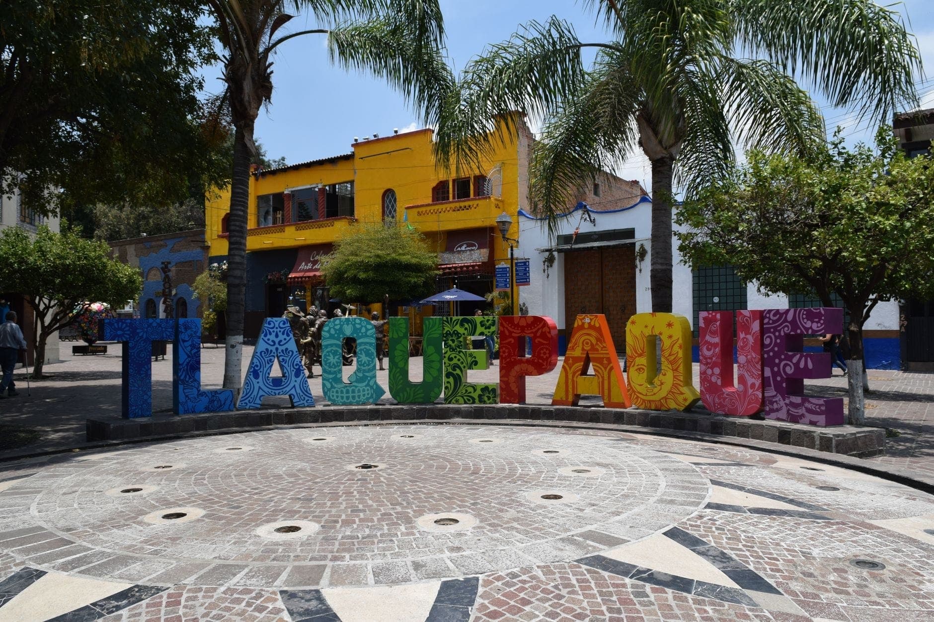 ป้าย Tlaquepaque สีสดท่ามกลางต้นปาล์ม ตึกสีเหลืองสดใส และลานที่คึกคัก ถ่ายทอดความมีชีวิตชีวาของ Andador Independencia