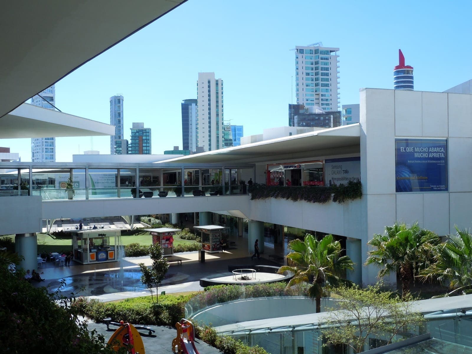 สถาปัตยกรรมสมัยใหม่และสวนที่เขียวชอุ่มที่ Andares Shopping Mall ในเมือง Zapopan พร้อมตึกระฟ้าสูงตระหง่านบนพื้นหลังท้องฟ้าสีฟ้าสดใส
