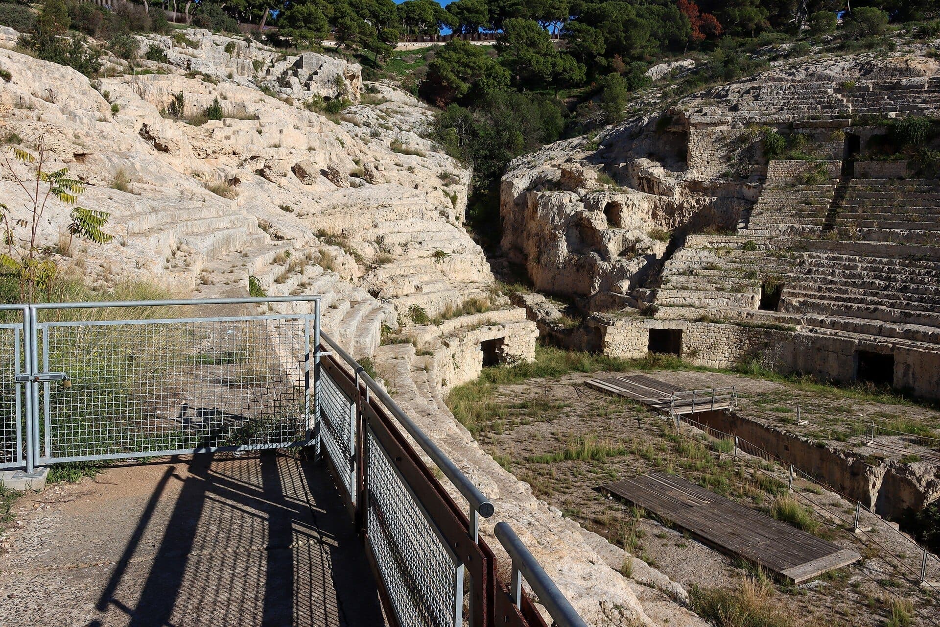 Vista del antiguo Anfiteatro Romano de Cagliari, con asientos de piedra desgastada tallados en una ladera y una puerta metálica cerrada en primer plano.