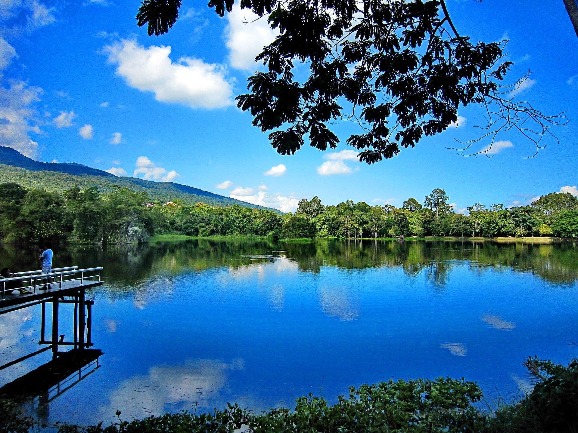 Der Ang Kaew Stausee an der Chiang Mai University, umgeben von üppigen Bäumen, klare Spiegelungen im See, Berge und blauer Himmel im Hintergrund.