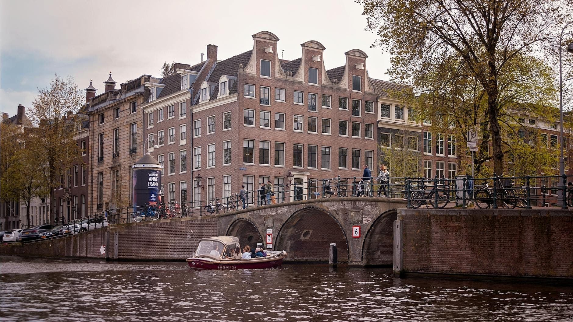 Weiter Blick auf das Anne-Frank-Haus und das Museum am Prinsengracht-Kanal in Amsterdam, mit einem Boot, das unter einer historischen Bogenbrücke hindurchfährt.