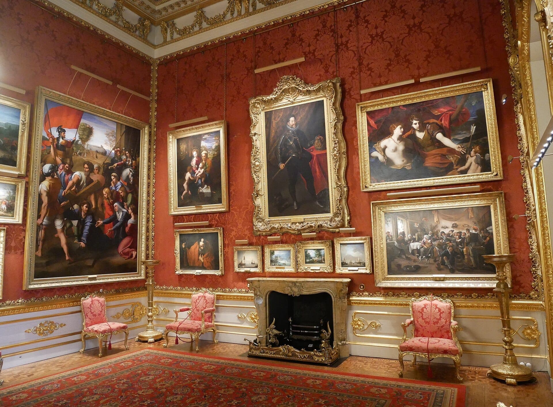 Wystawna galeria sztuki w Apsley House z obrazami w złotych ramach, czerwoną tapetą we wzory, ozdobnym kominkiem i antycznymi krzesłami tapicerowanymi w kolorze różowym.
