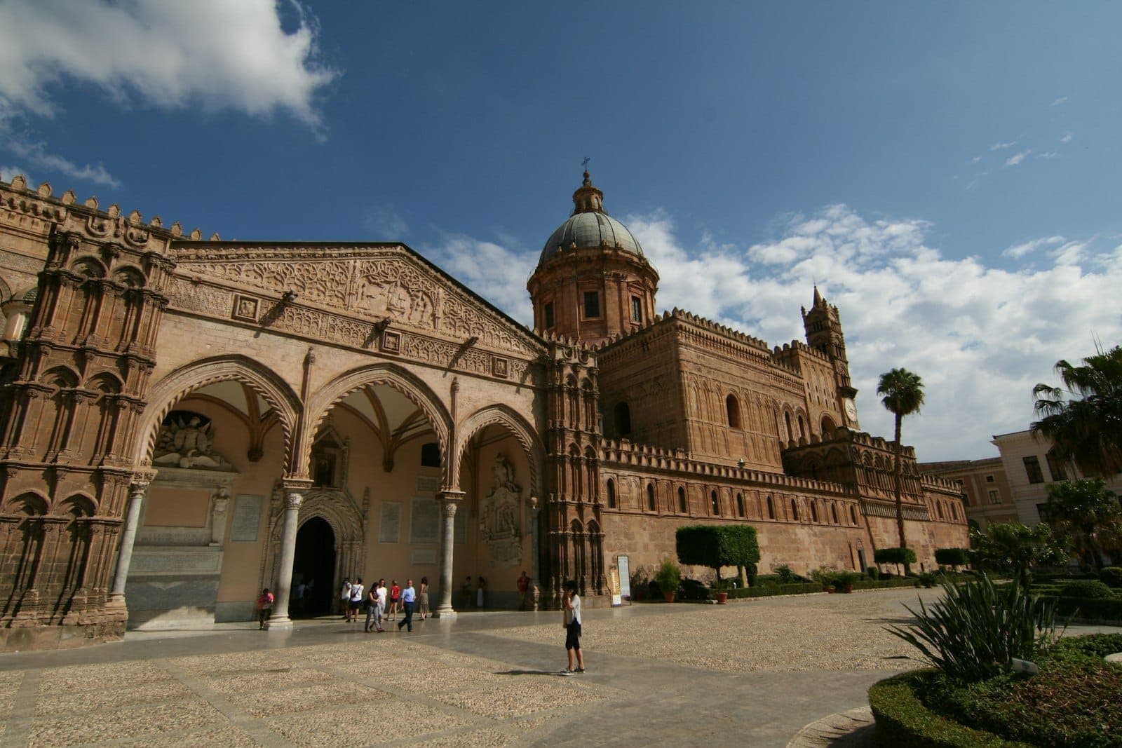 Pemandangan luas Katedral Palermo di Sisilia yang menampilkan ciri arsitektur Arab-Norman dengan lengkungan, ukiran batu, kubah, dan orang-orang di luar.