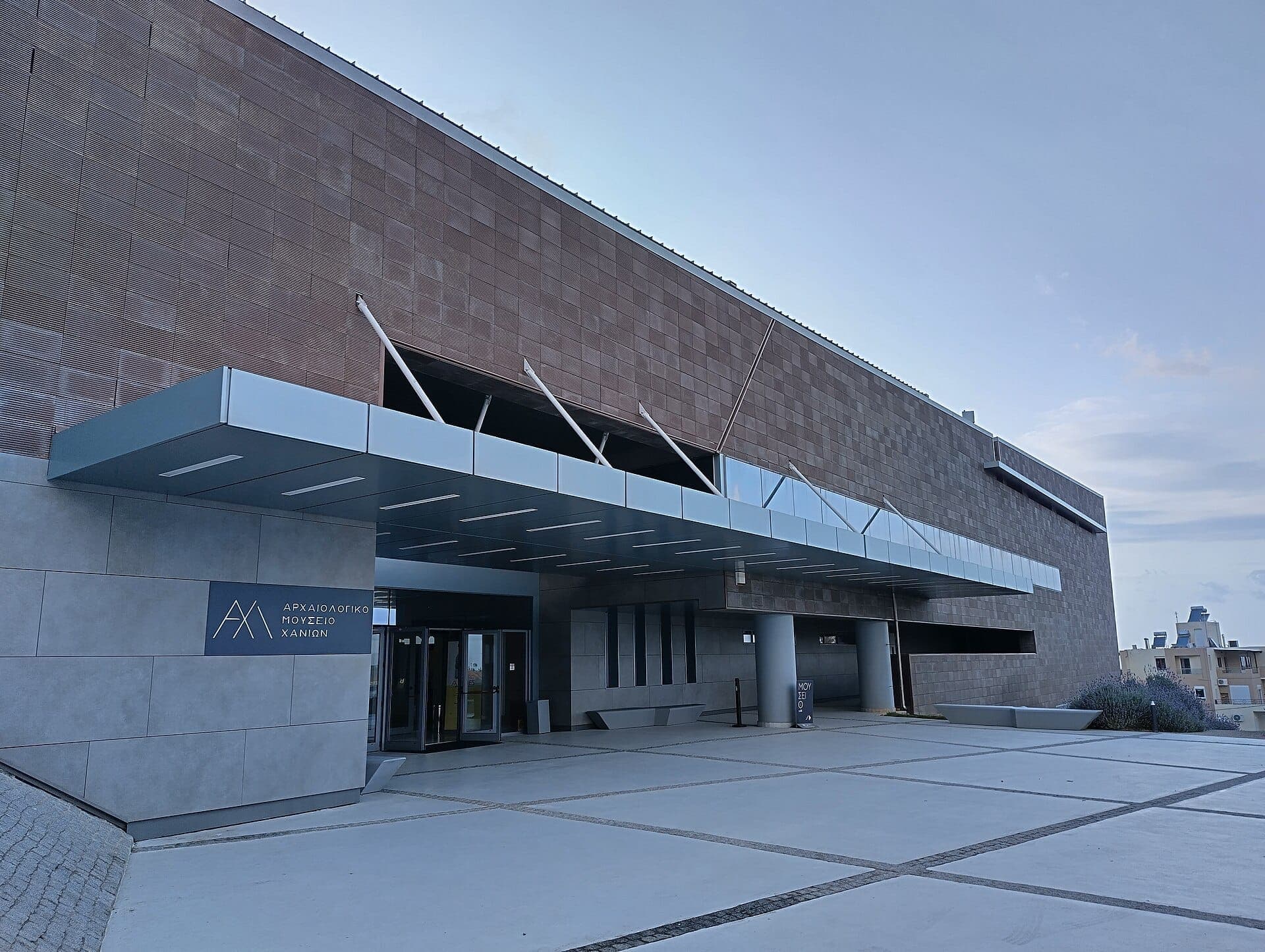Modernes Äußeres des Archäologischen Museums Chania mit geometrischen Linien, großem Glasvordach und strukturierter Ziegelfassade im sanften Tageslicht.