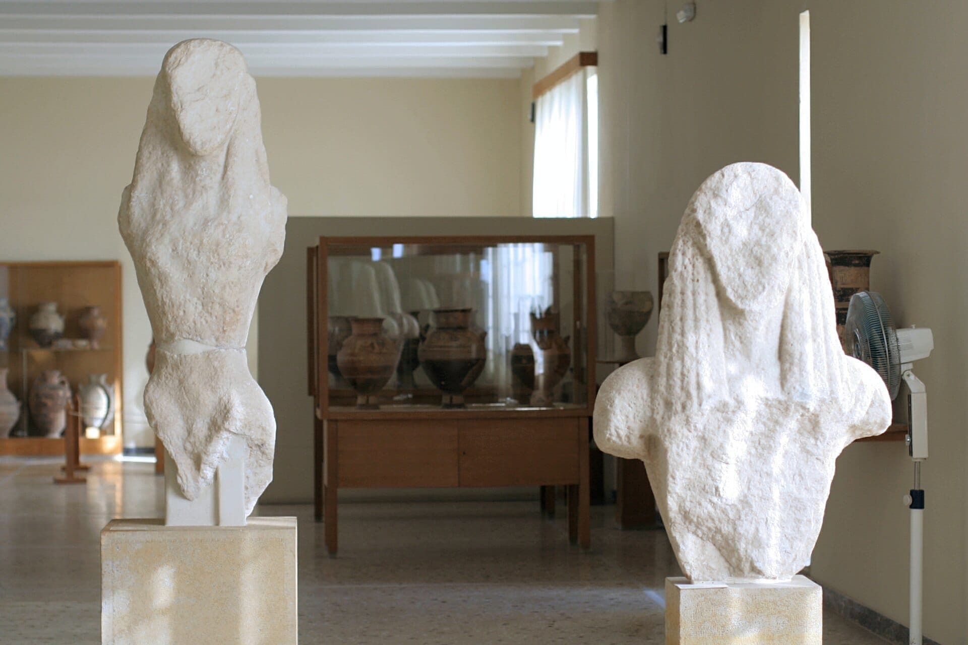 Zwei antike Marmorstatuen auf Sockeln in einer hellen Galerie des Archäologischen Museums von Thera, mit Terrakottavasen in Glasvitrinen im Hintergrund.