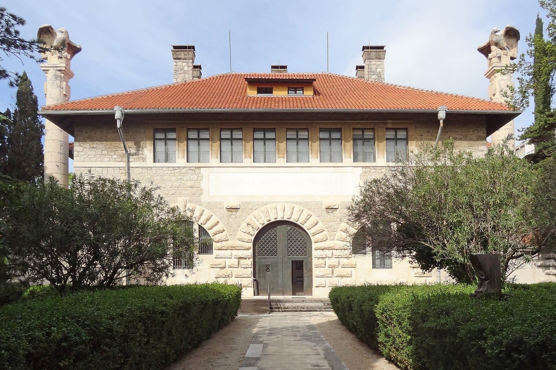 Facciata del Museo Archeologico di Split con muri in pietra, tetto in tegole rosse, ingresso ad arco e verde simmetrico sotto un cielo luminoso.