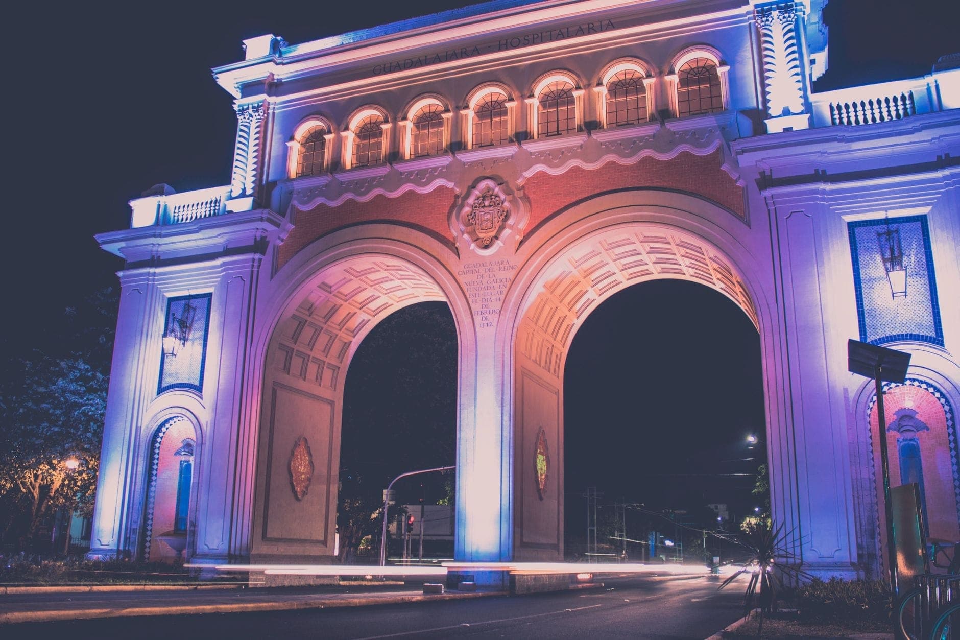 วิวกลางคืนของอนุสรณ์ Arcos de Guadalajara ที่ประดับไฟม่วงและน้ำเงิน เห็นซุ้มทั้งสองข้ามถนนใหญ่โล่งเงียบ