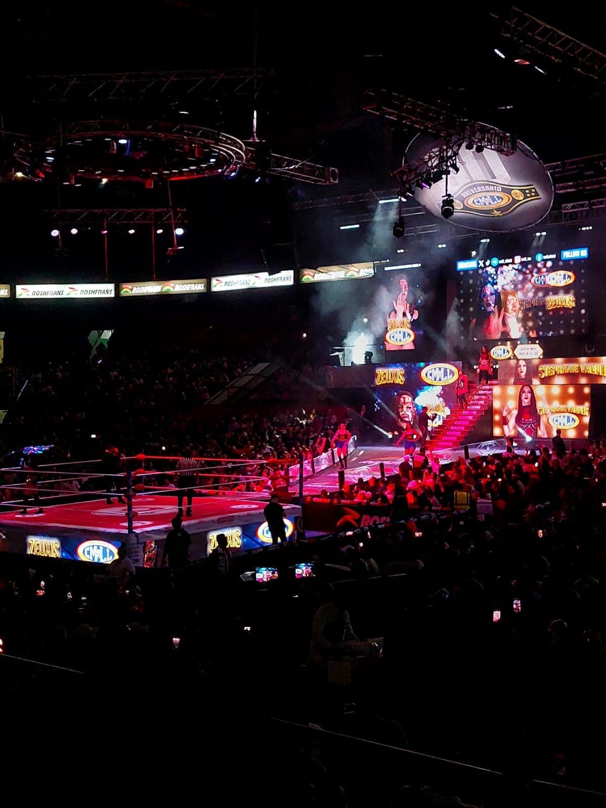 นักมวยปล้ำเดินเข้าสู่สังเวียนที่สว่างไสวของ Arena México ขณะที่ฝูงชนที่โห่ร้องเต็มอาคารที่มืดมิด พร้อมจอภาพสดใสและสปอตไลต์เหนือศีรษะ