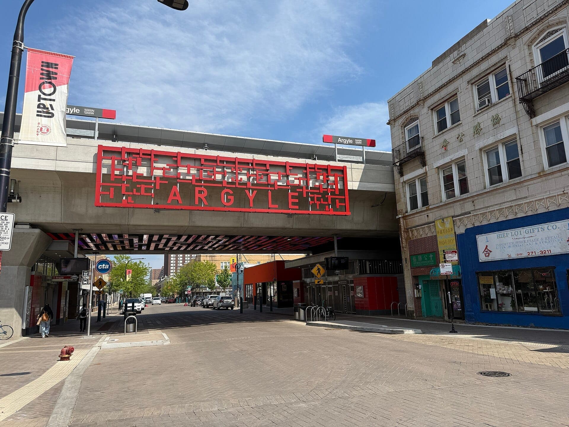 Street-level view of Argyle Street's viaduct and surrounding shops in Chicago's Uptown, featuring colorful signage and clear blue sky.