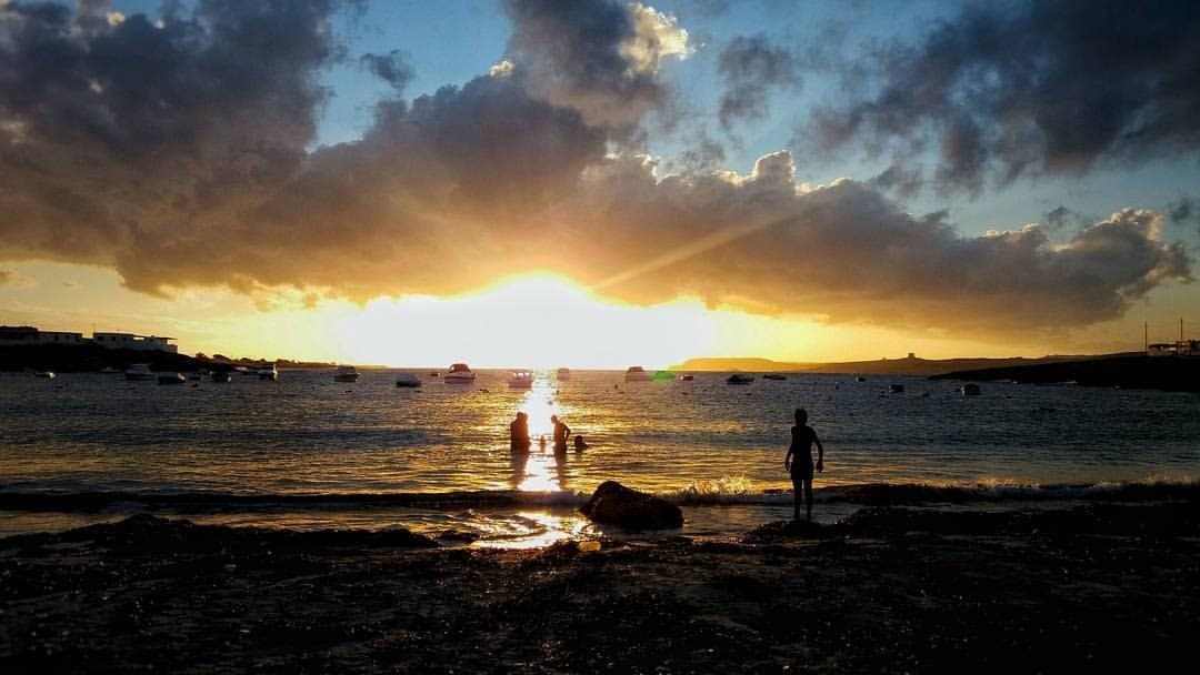 Sonnenuntergang an der Armier Bay, Malta, mit Silhouetten von Menschen am Strand, Booten auf ruhigem Wasser und dramatischen Wolken am Himmel.