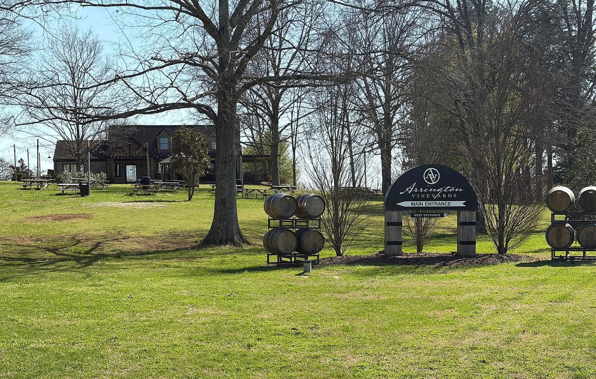 Arrington Vineyards main entrance sign, wine barrels, and a rustic building set on grassy, tree-dotted grounds under bright daylight.