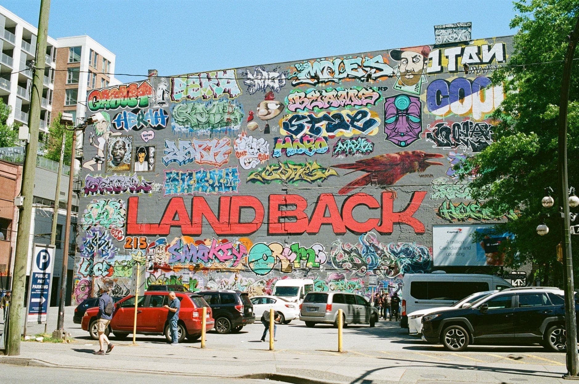 จิตรกรรมฝาผนังกลางแจ้งขนาดใหญ่พร้อมกราฟฟิตีสีสันสดใสและข้อความ 'Land Back' ที่โดดเด่นบนกำแพงในย่านเมืองของ Vancouver มีรถจอดและผู้คนอยู่ใกล้เคียง