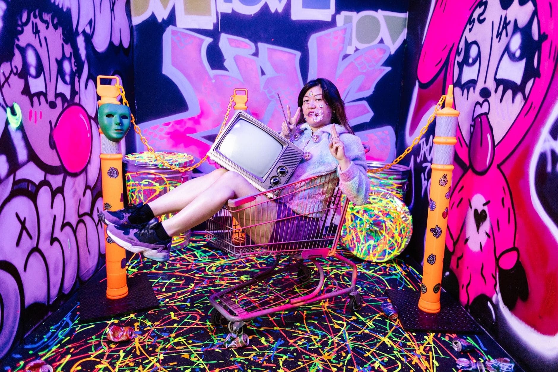Eine junge Frau posiert in einem Einkaufswagen mit einem Retro-Fernseher im Art in Paradise Chiang Mai, umgeben von lebhaften Neon-Graffiti und Kunstwerken.