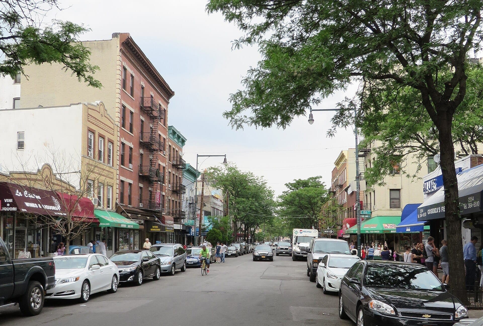 วิวถนน Arthur Avenue ใน The Bronx มีอาคารอิฐเก่าแก่ รถยนต์จอดอยู่ ผ้าใบหน้าร้าน และต้นไม้ใบเขียวในวันที่อากาศสดใส