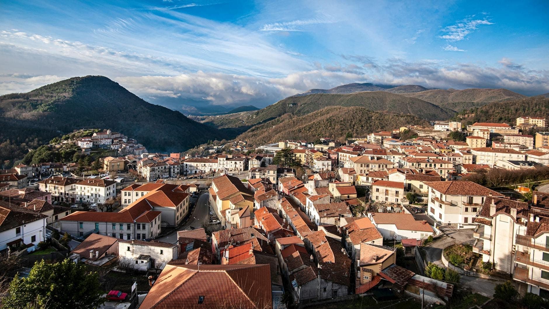 Vista panorámica de un pueblo sardo de montaña con tejados de tejas rojas entre colinas boscosas y cumbres imponentes bajo un cielo azul con nubes.