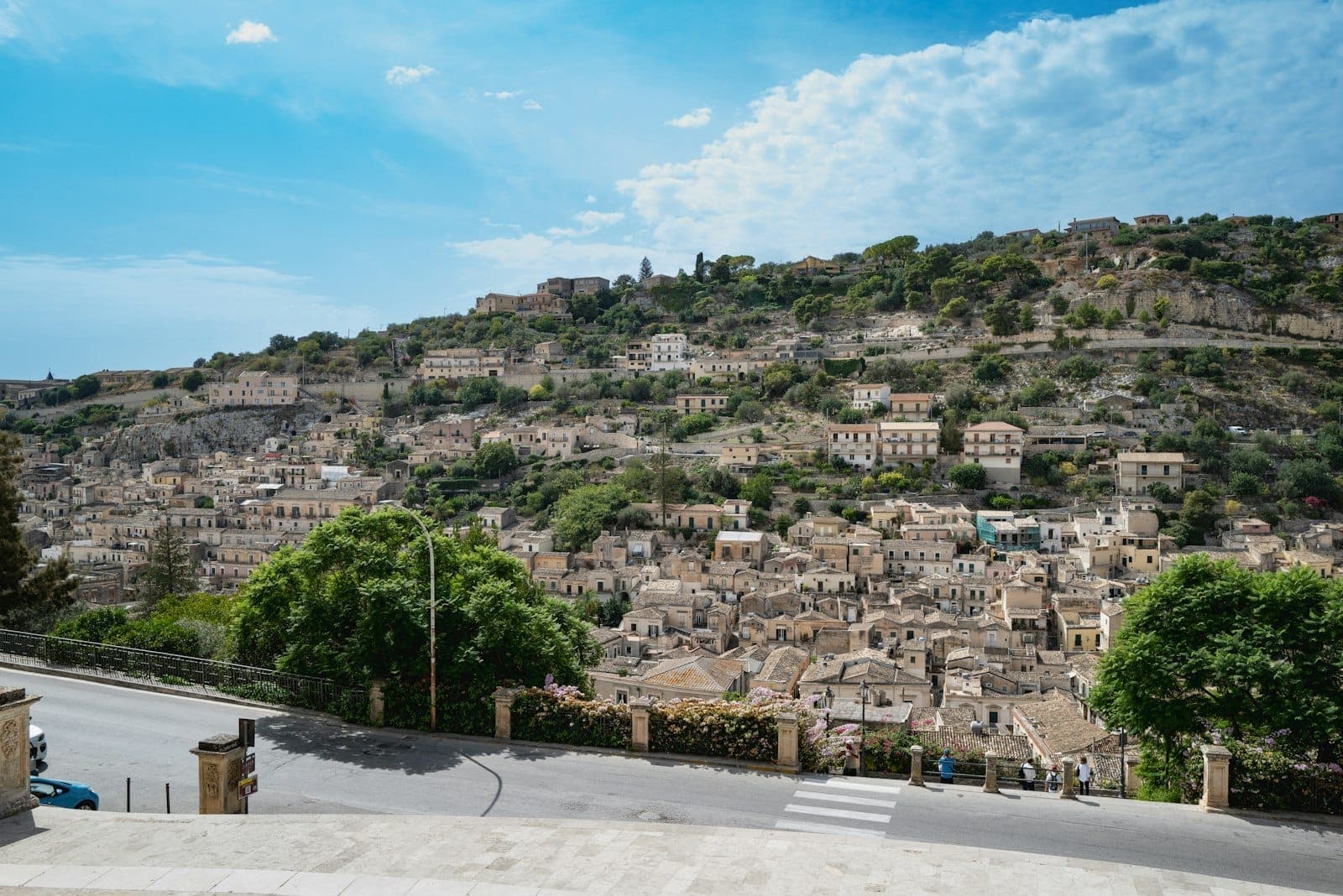 Pemandangan panoramik kota barok Modica di Sisilia, dengan bangunan batu yang padat di lereng hijau di bawah langit cerah.